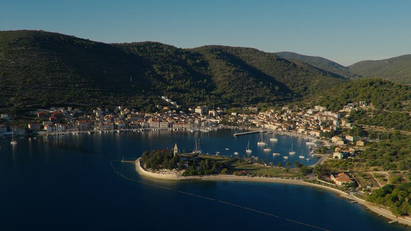 Aerial view of a harbour town on green hills, with sailboats anchored in the bay.