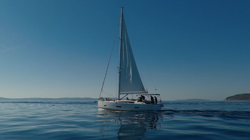 A white sailboat with its sails up on calm blue water, with people standing in the cockpit.