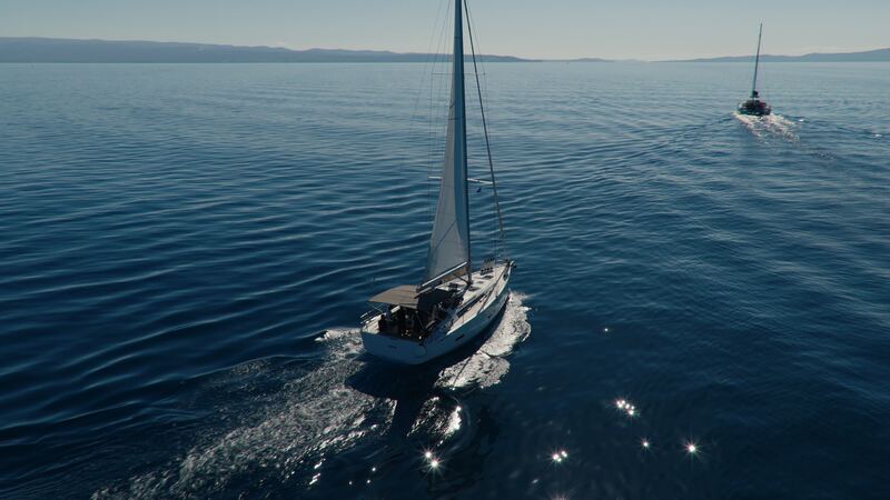 A sailboat under main and headsail motors across rippled blue water, seen from above and behind, with a second boat ahead.