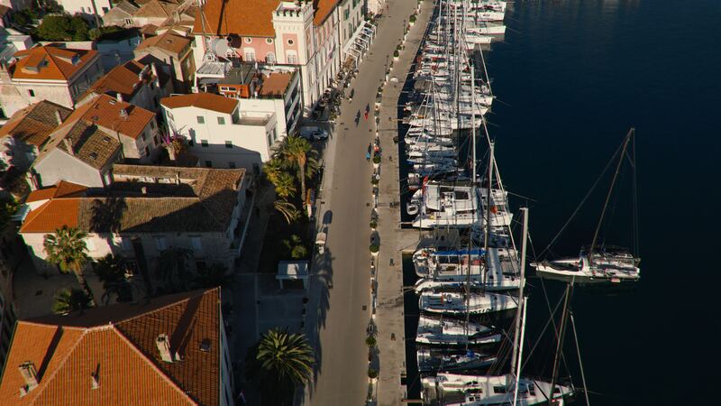 Sailboats moored stern-to along a harbour quay, seen from above, beside a street with palm trees and red-roofed houses.